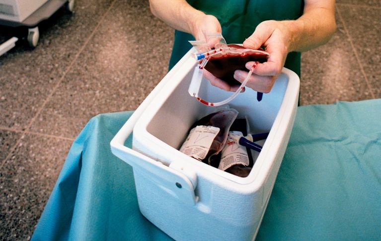 Ein Mann in Operationskleidung nimmt in einem Operationssaal eine Blutkonserve aus einer Kühlbox mit mehreren Blutbeuteln